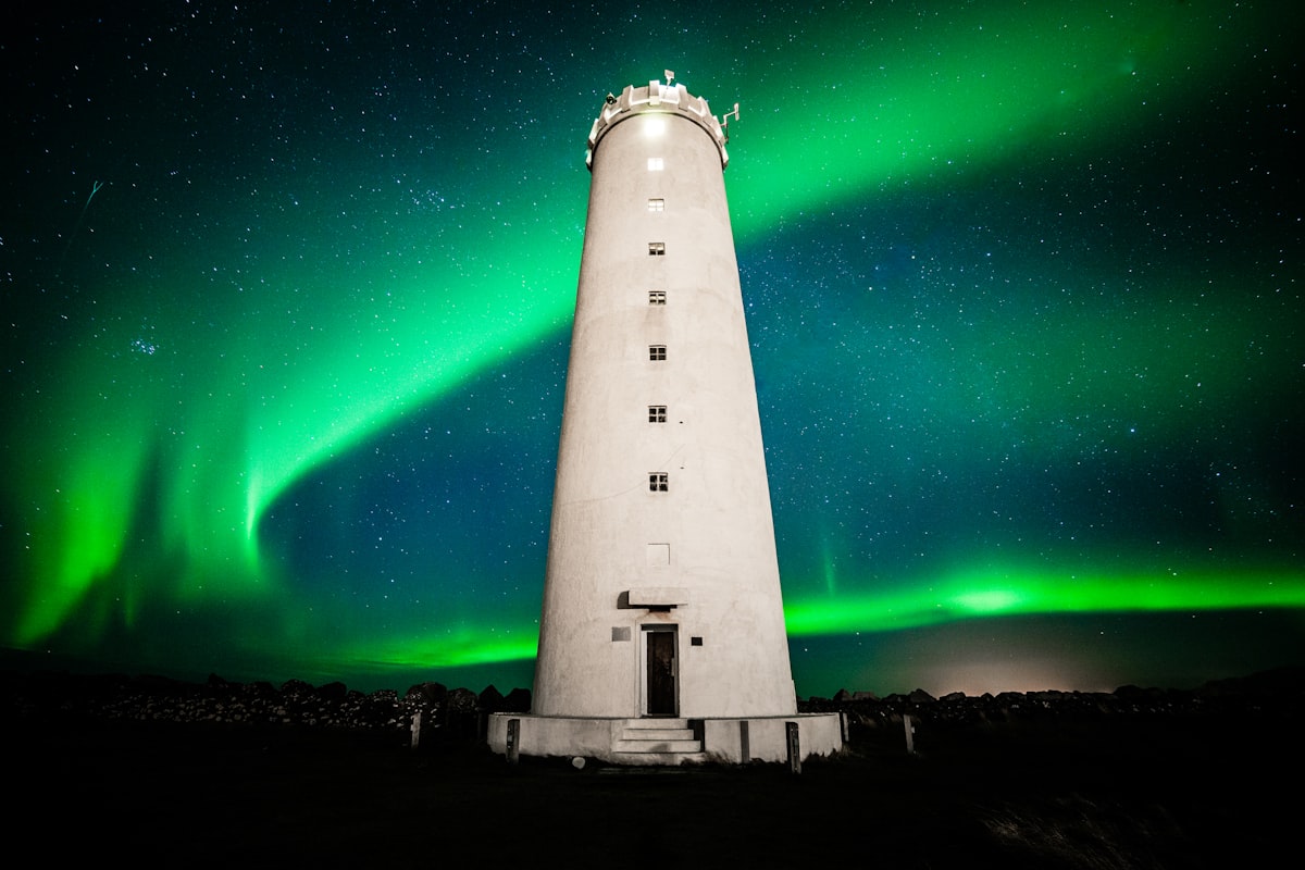 Green northern lights arc over Seltjarnarnes lighthouse in Iceland, reflected in the calm sea below