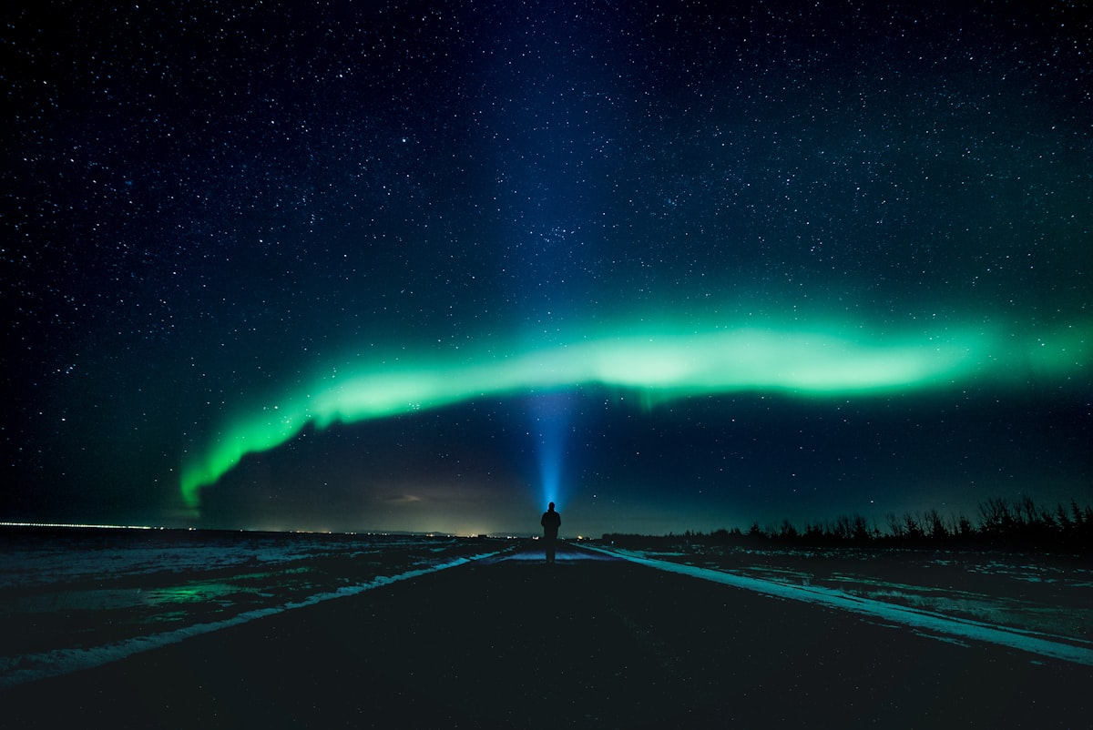 A green aurora arc stretched across a black, star-filled sky above a straight Icelandic road, with a single figure standing in the middle of it. The density of visible stars is what a genuinely moon-free night looks like.