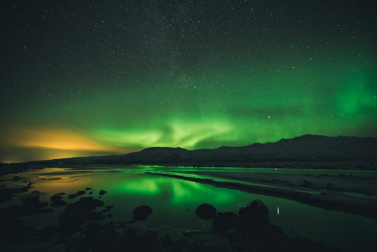 Long-exposure aurora over a still lake near a mountain in Þórsmörk, Iceland, with a hint of light pollution from a town to the left.