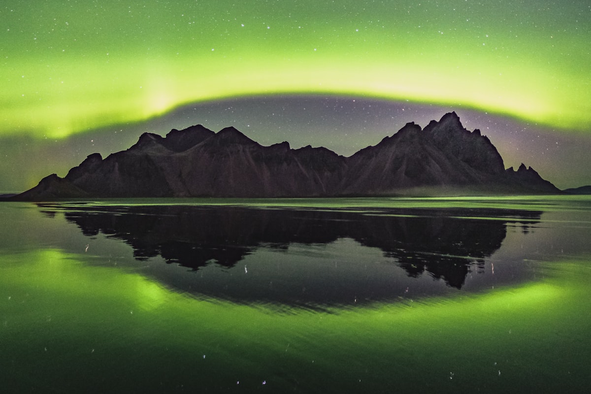 A vivid green aurora arc curving over the jagged peaks of Vestrahorn in southeast Iceland, mirrored in the still water of Stokksnes lagoon.