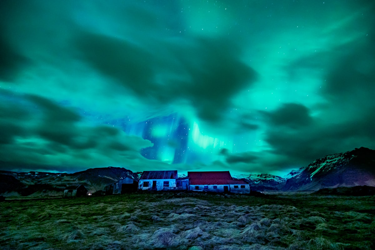 A lone Icelandic farmhouse standing in open grassland under a bright green aurora, with no other light anywhere in the frame.