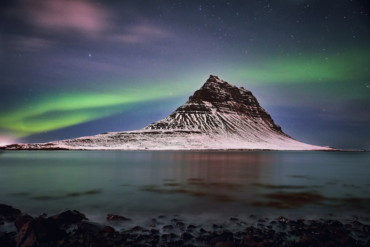 A snow-capped mountain stands beneath a green aurora arc on a clear winter night in Iceland.