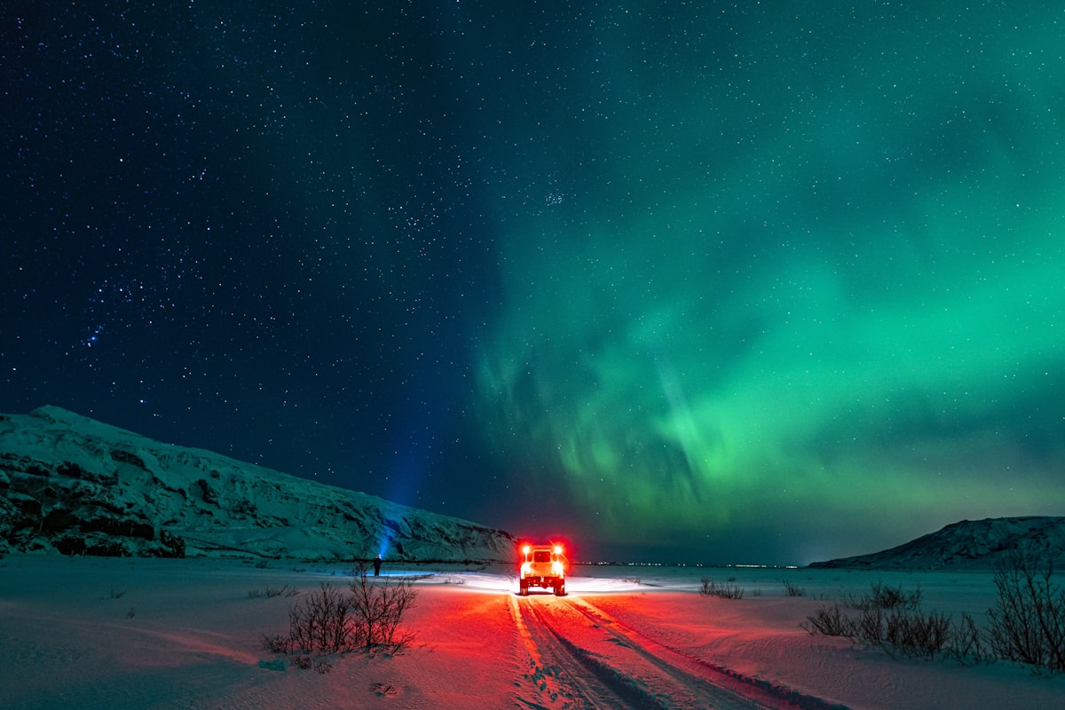 A car driving along a snow-covered Icelandic road at night under a green aurora, headlights lighting the way ahead.