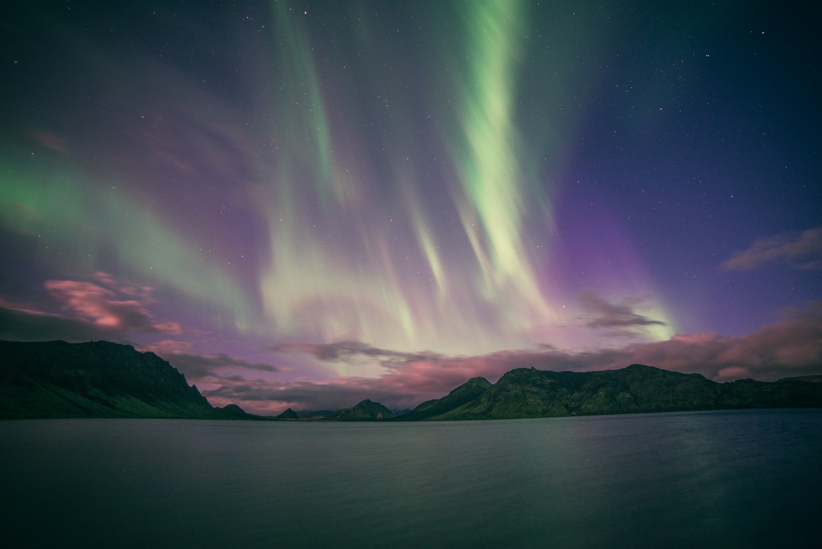 Green and violet aurora curtains rising over Álftavatn lake in the Icelandic highlands, with snow-free green hills and pink-lit clouds low on the horizon: an early-season display in a sky that is not yet fully dark.