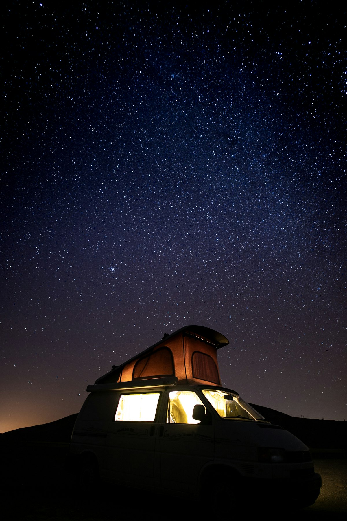 A camper van beneath a dense field of stars at night with its lights off, the kind of dark sky an aurora chaser is trying to park under.