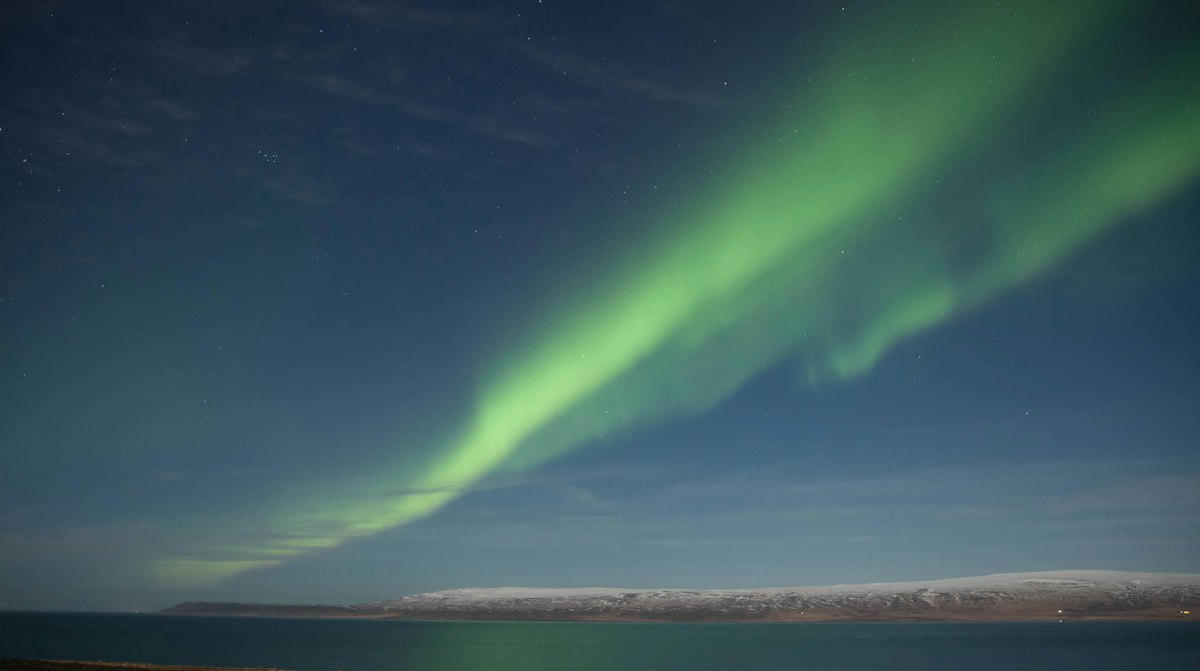 A green aurora arc sweeping across a deep blue sky over a fjord near Búðardalur in west Iceland, with snow on the hills across the water.