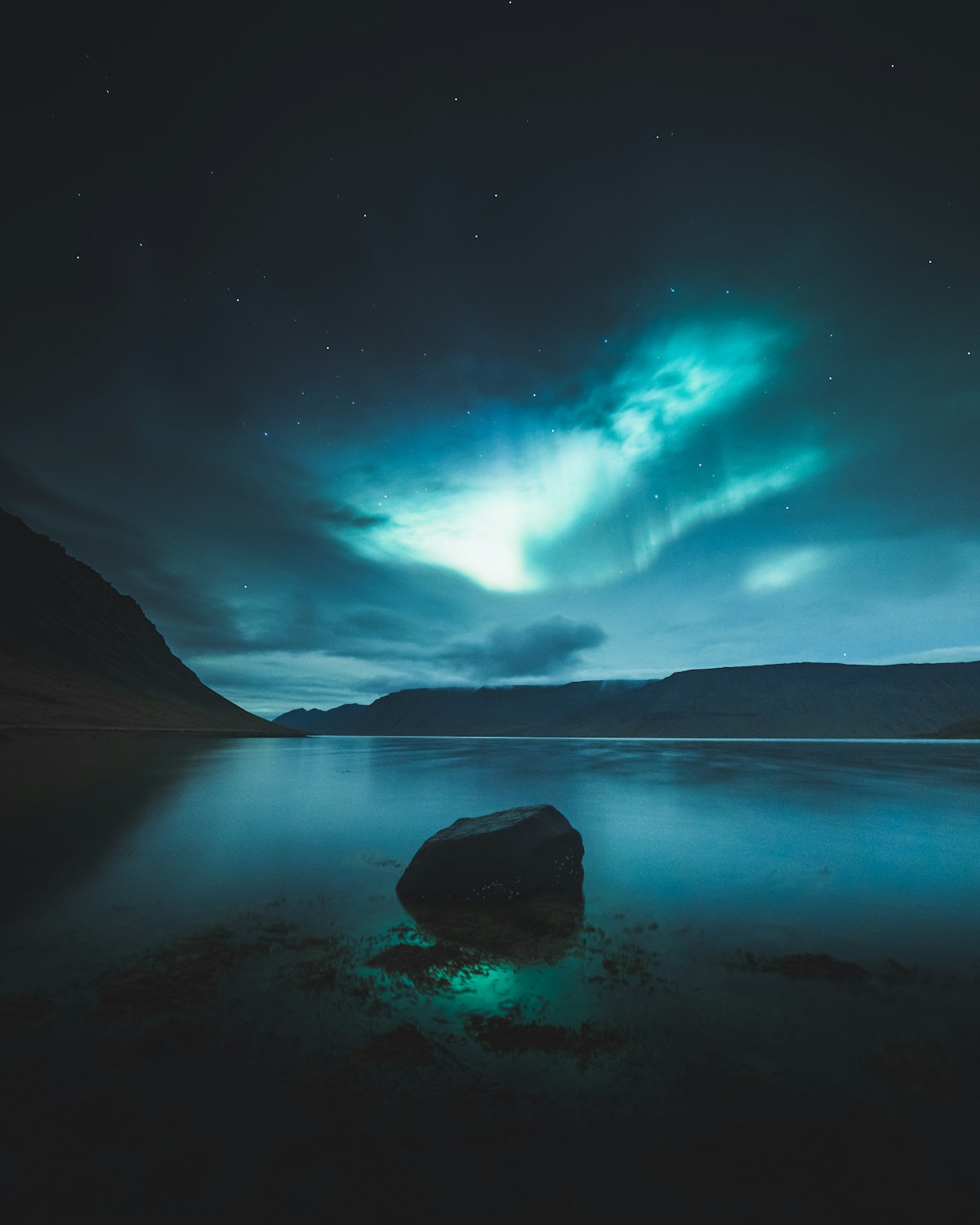 The northern lights arc over a dark lake and low hills in the Icelandic wilderness, with a band of stars visible above the horizon.