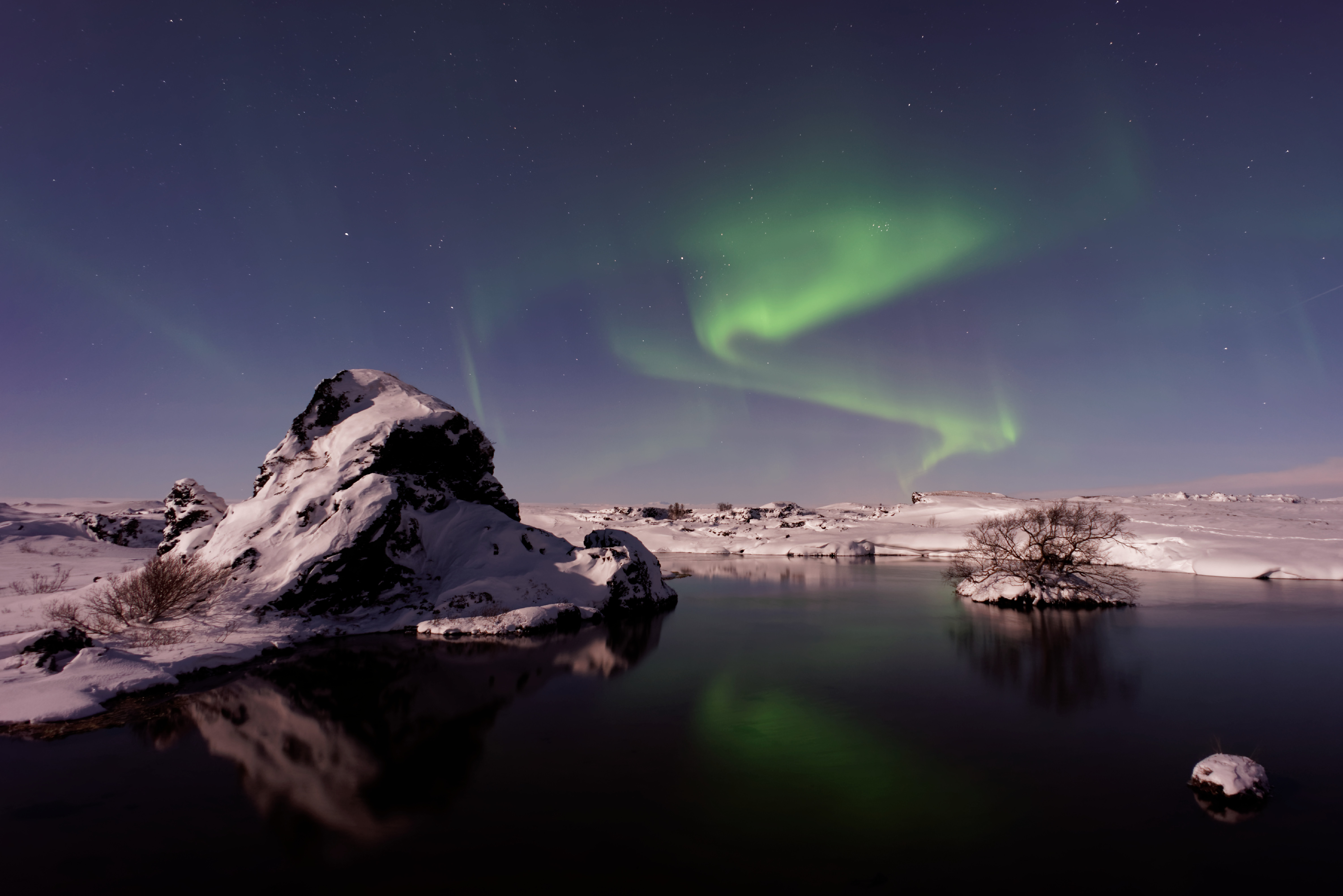 Green northern lights swirling above a frozen Icelandic lake at night, reflected in the still water with snow-covered lava rocks in the foreground