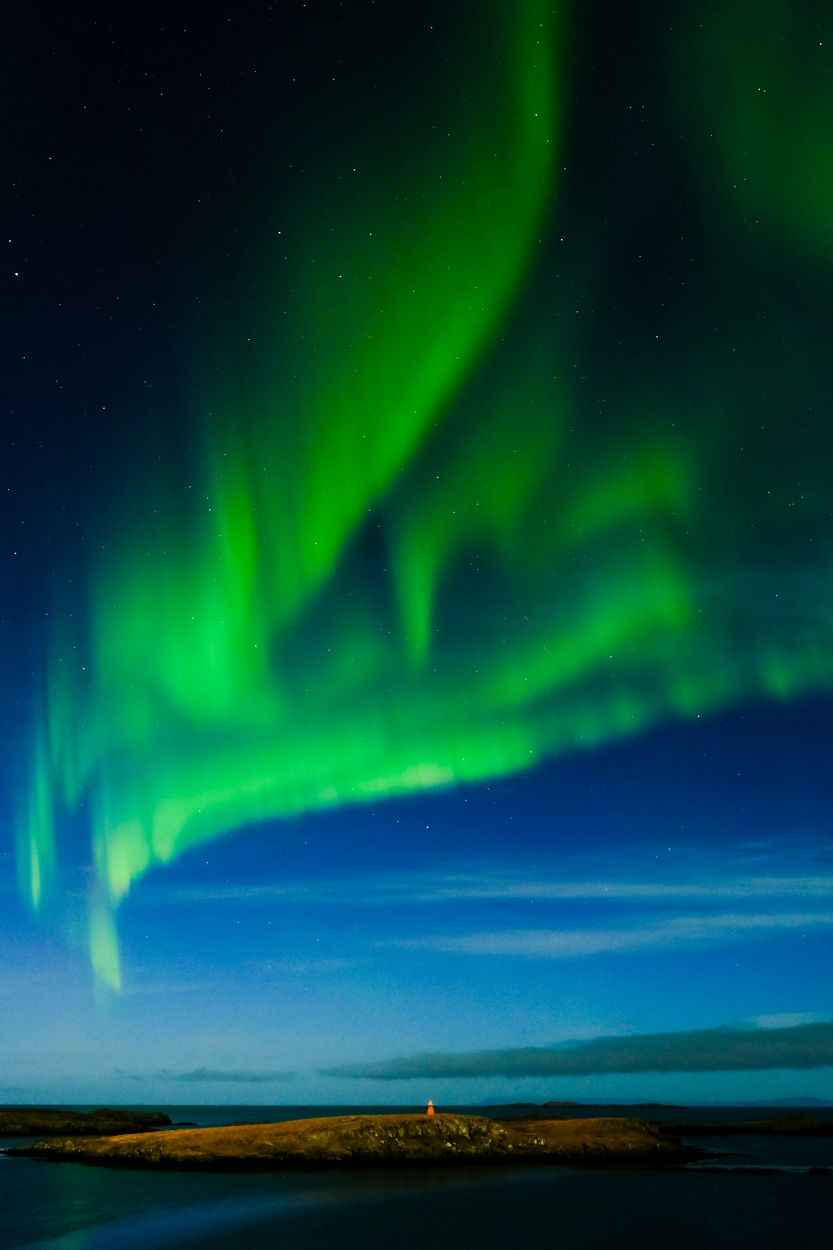 Vibrant green aurora borealis dances across a dark sky above a coastal Icelandic landscape, with the silhouette of low hills against the horizon.