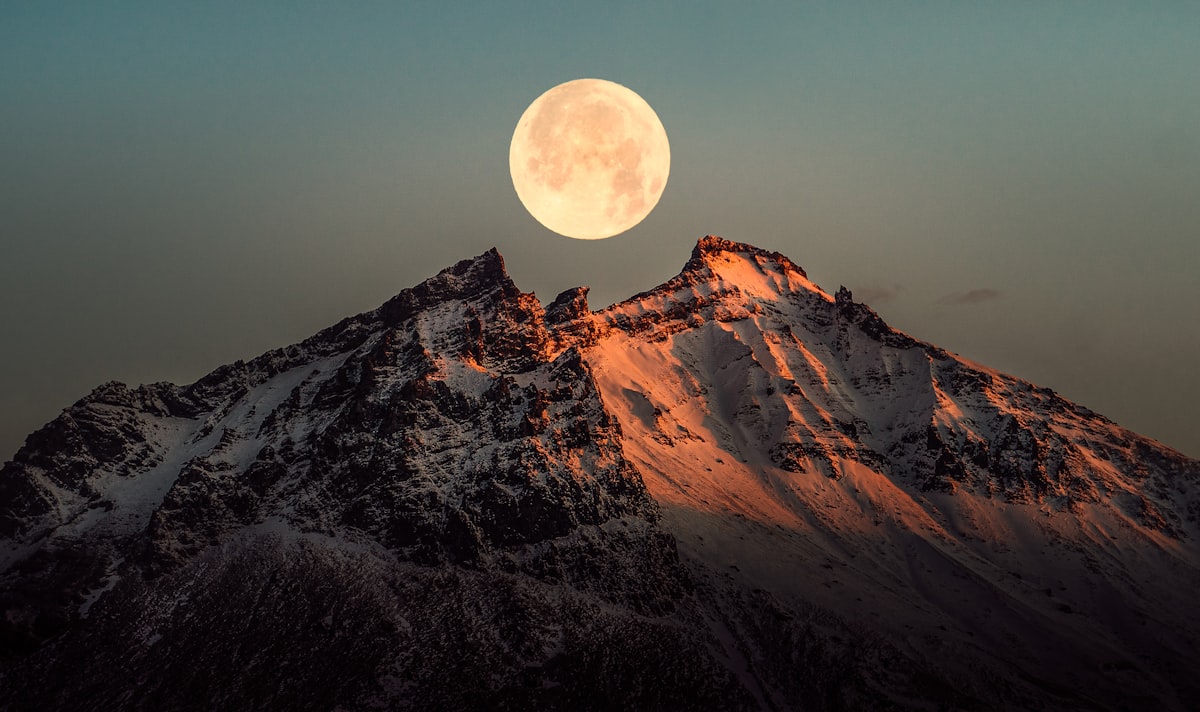 A bright full moon rising directly above a snow-covered mountain ridge against a deep blue twilight sky.