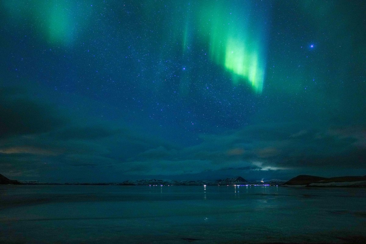 Une aurore verte suspendue au-dessus du lac Mývatn la nuit, avec les lumières éparses d'un hameau le long de la rive opposée