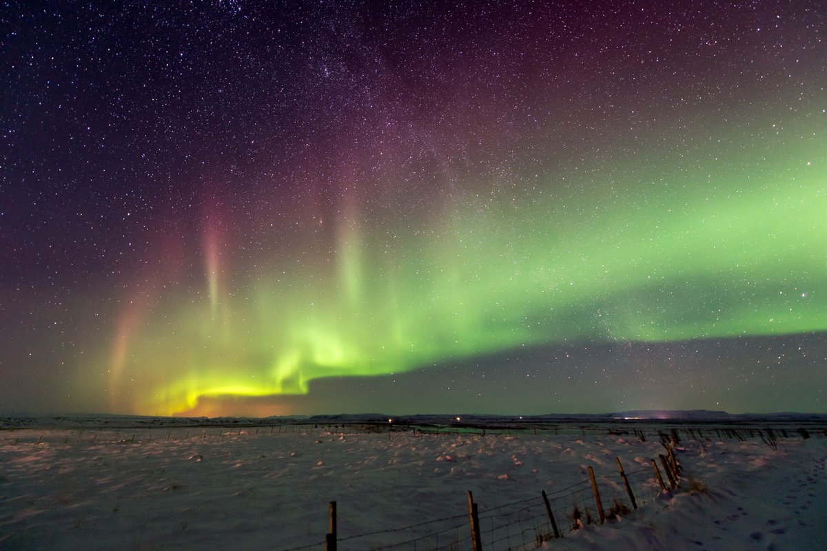 Un large arc d'aurore vert et violet remplit le ciel au-dessus de champs enneigés et d'une clôture dans le sud de l'Islande