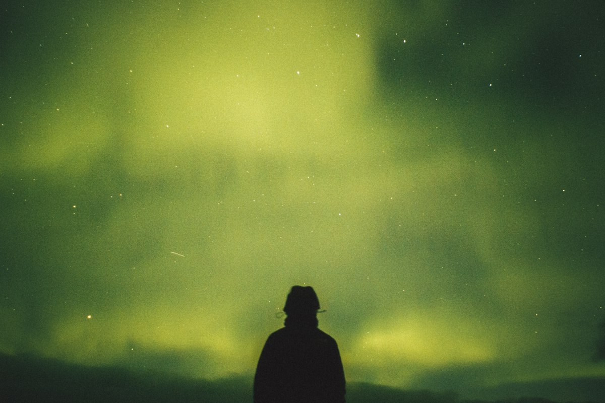 A lone figure stands looking up at green northern lights across the night sky in Iceland, the way a tour group stands once the bus has stopped.