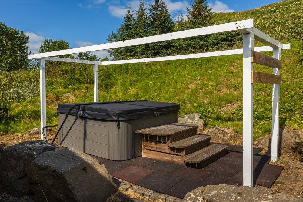 A covered hot tub with wooden steps under a white pergola, set into a grassy bank at Hestheimar
