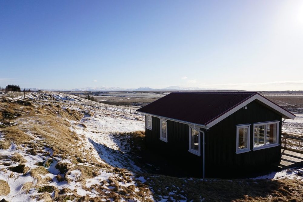 A dark-painted Hestheimar cottage on a hillside dusted with snow, under a clear winter sky