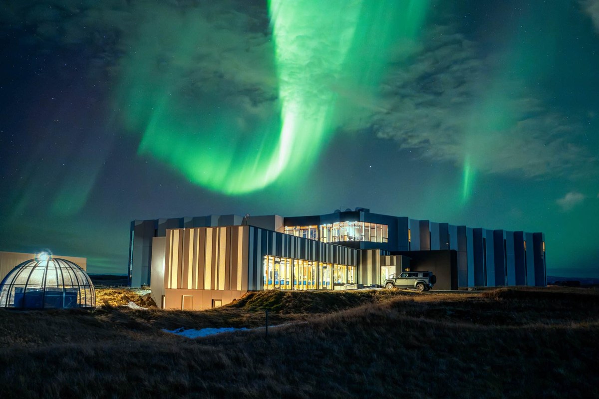 Landhotel lit up at night under a green aurora curtain, with its glass viewing dome on the grass alongside